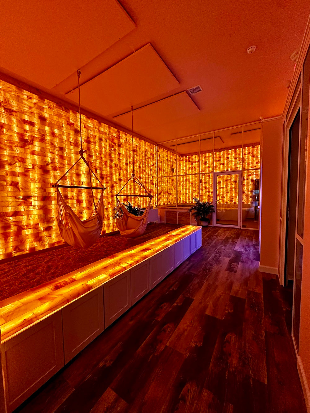 A warmly lit interior room with Himalayan salt walls creates a soft glow. Two hammock chairs hang over crushed Himalayan salt.  A long, illuminated Himalayan salt bench runs along the middle of the room, contrasting with the dark wooden floor. The atmosphere is serene and inviting, evoking a sense of relaxation.