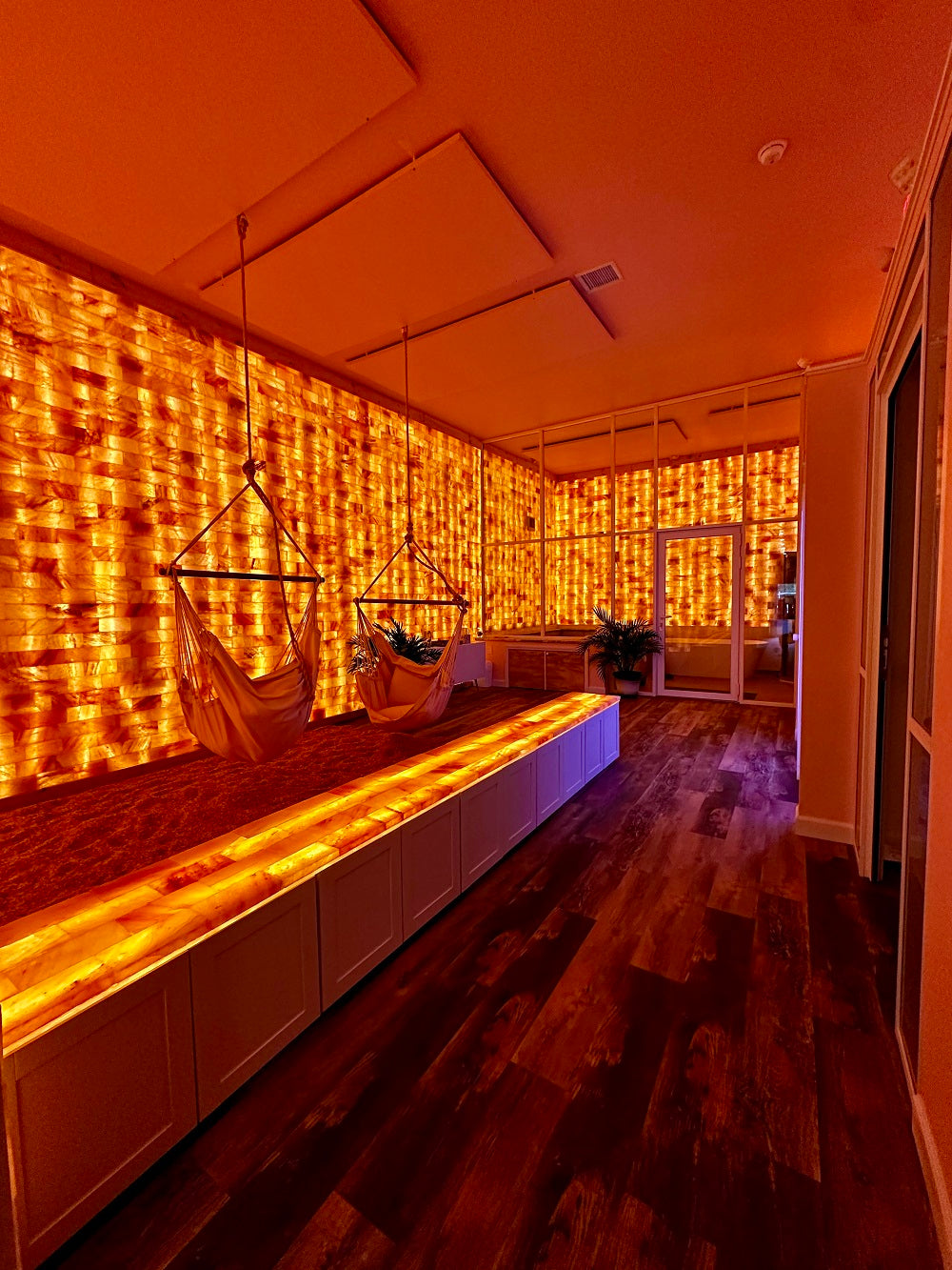 A warmly lit interior room with Himalayan salt walls creates a soft glow. Two hammock chairs hang over crushed Himalayan salt. A long, illuminated Himalayan salt bench runs along the middle of the room, contrasting with the dark wooden floor. The atmosphere is serene and inviting, evoking a sense of relaxation.